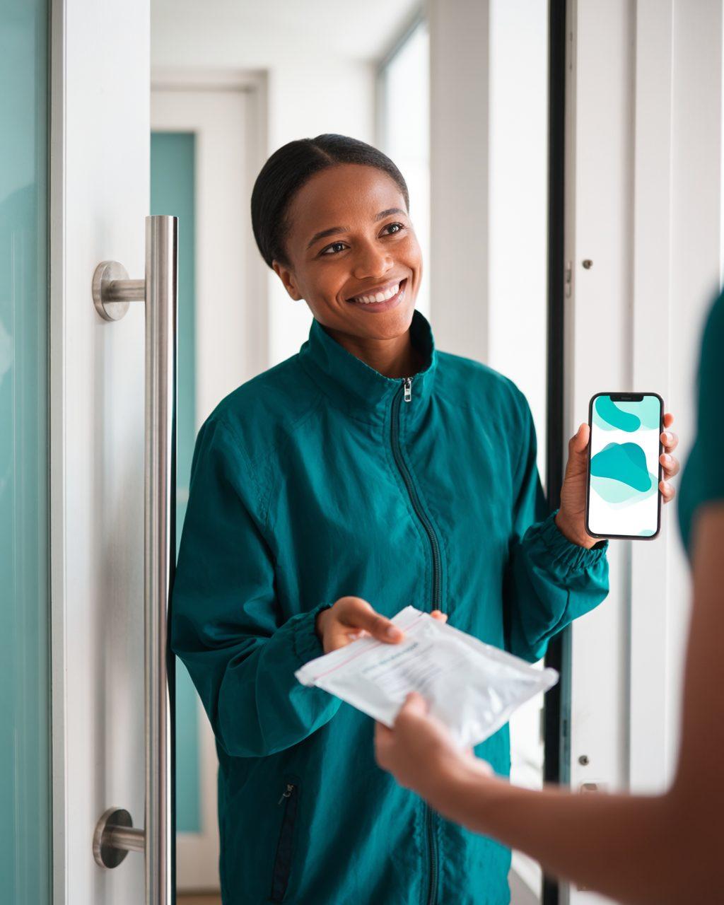 Entregadora sorridente entrega um pacote lacrado da farmácia na porta de um apartamento, com o pedido aberto no celular.