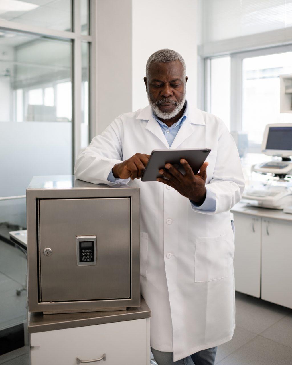 A pharmacist reviews records on a tablet next to the locked controlled-substances cabinet with a keypad.